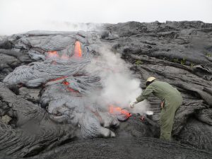 Amidst steam created by rain falling on the hot lava, another HVO geologist uses a rock hammer to collect a sample of the active flow, Wednesday. USGS/HVO photo.