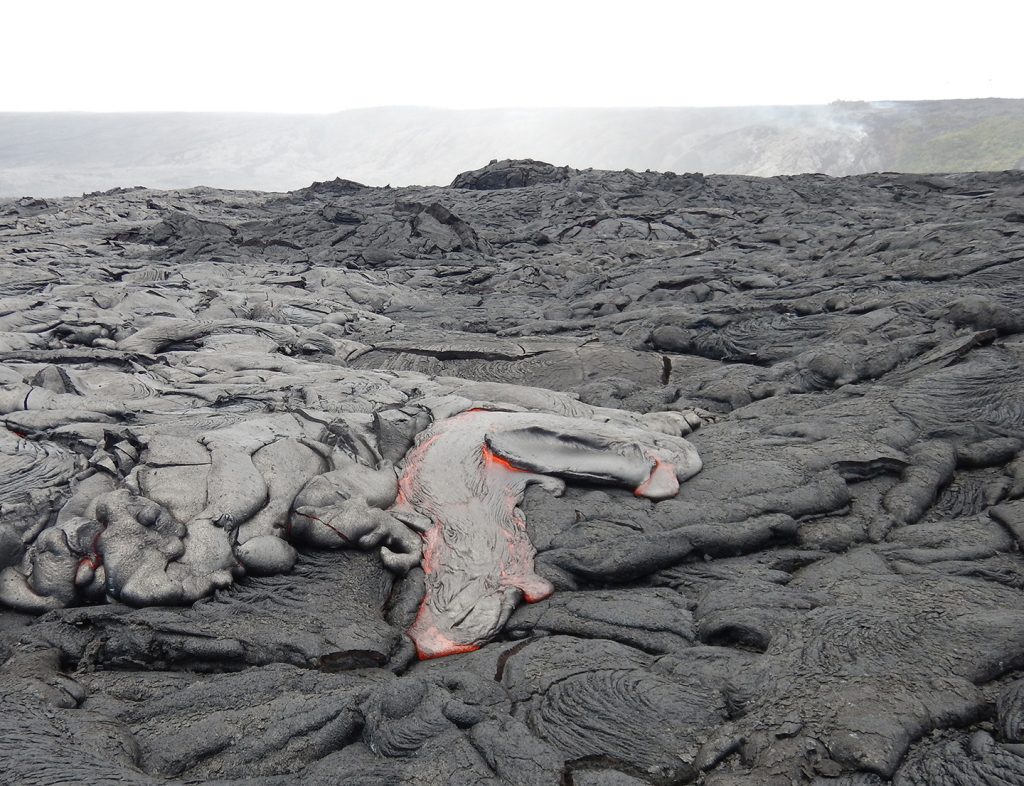 The active lava flow continues to creep across the coastal plain on Kīlauea’s south flank. Breakouts of pāhoehoe (foreground) are fed by lava traveling through lava tubes from the Pu‘u ‘Ō‘ō vent and down Pulama pali (top background). USGS photo.